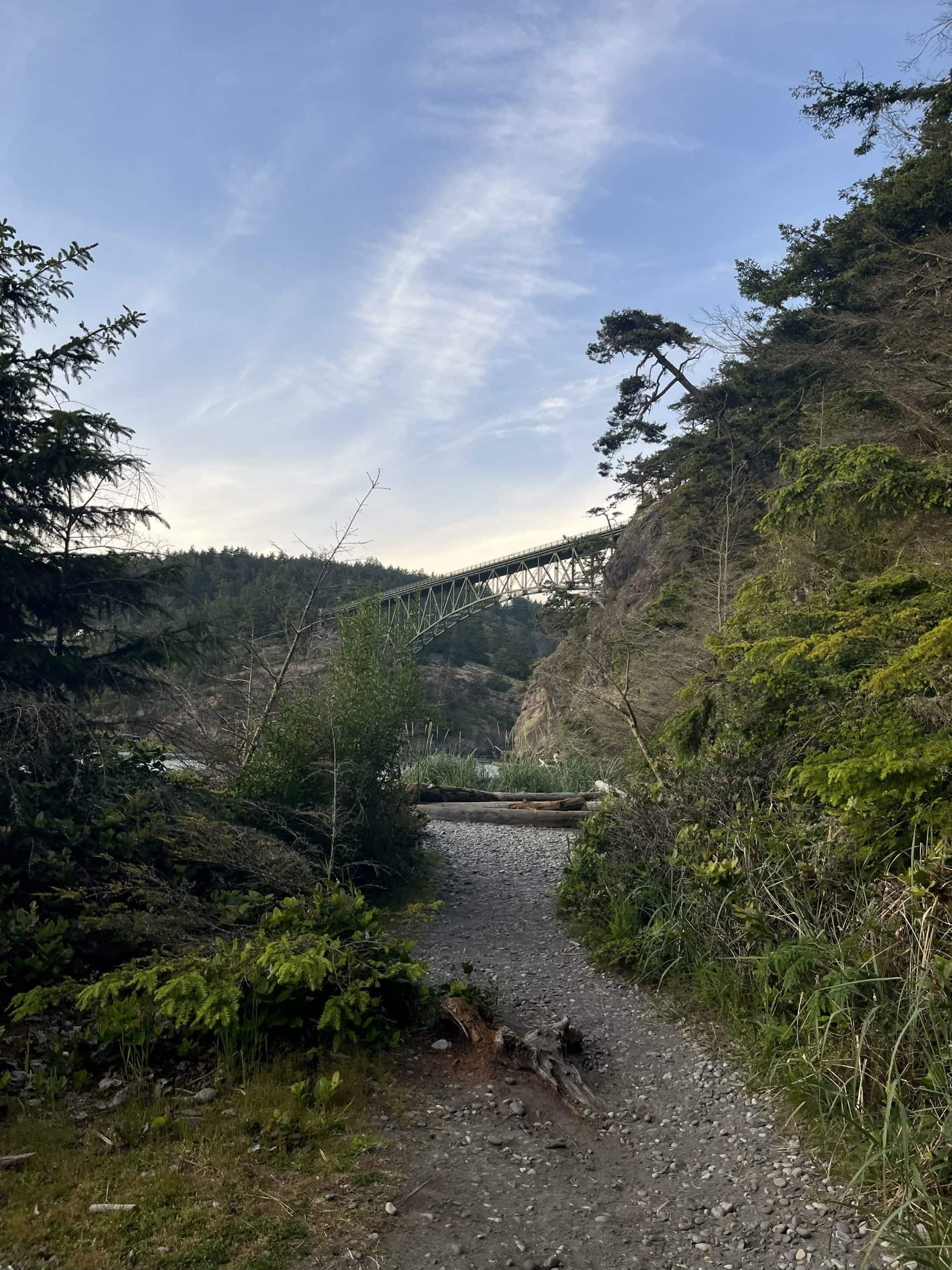 Trail leading through forest to Deception Pass Bridge on Whidbey Island, Washington, with cliffs and blue sky above.